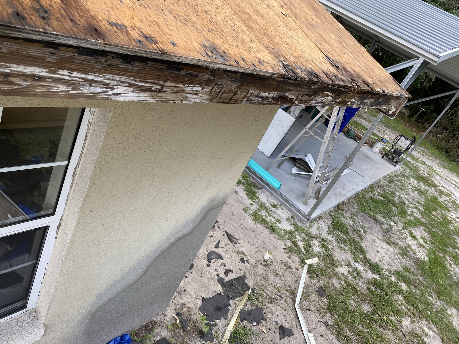 Rotted timber fascia and soffit exposed above a stucco wall, with a ladder resting against the eave